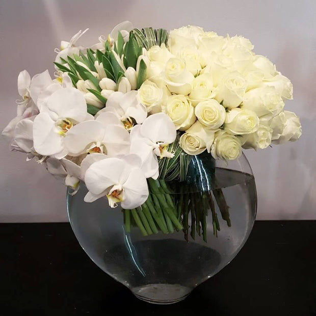 White flowers is displayed in a tight, modern design. Flowers in this design could include Peony, Roses, Amaryllis, Calla, Tulips, and White Cascading Orchids