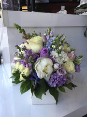 Lavender Lisianthus, Lavender Roses, White Roses, White Peony, Blush Snapdragon or Blush Hyacinth, Hints of Baby Blue Dalphinium or Hyacinth, and a mix of garden style greenery. Designed in a white vase.