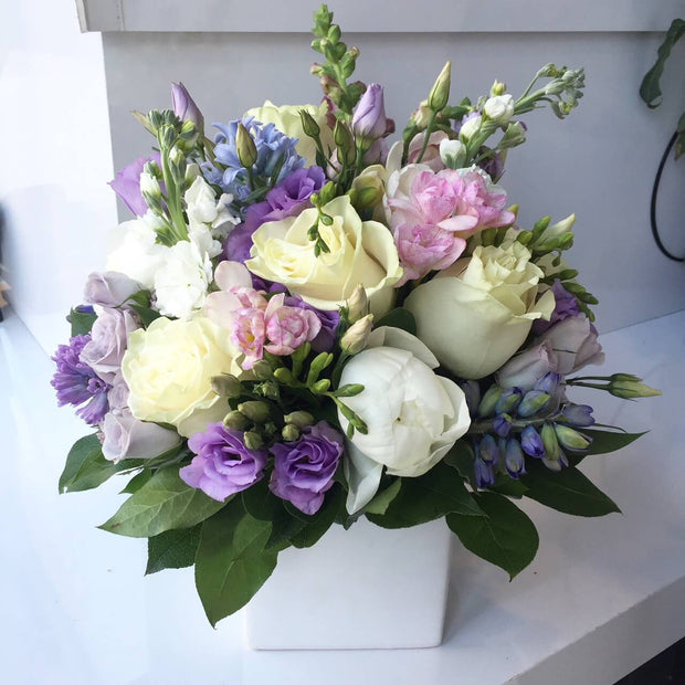 Lavender Lisianthus, Lavender Roses, White Roses, White Peony, Blush Snapdragon or Blush Hyacinth, Hints of Baby Blue Dalphinium or Hyacinth, and a mix of garden style greenery. Designed in a white vase.