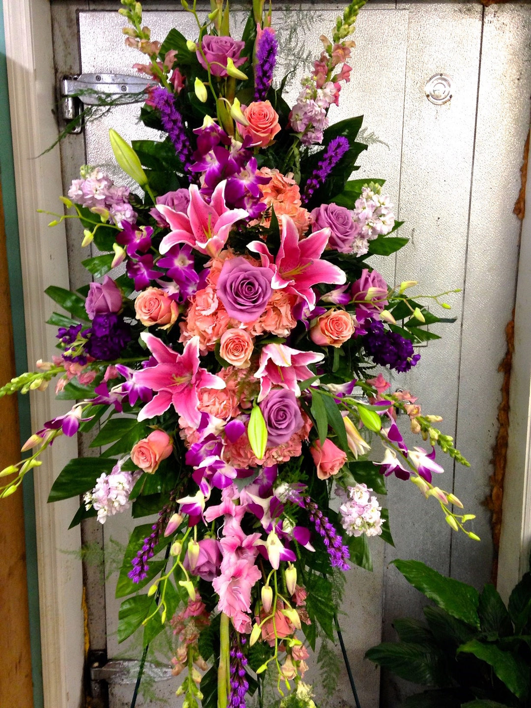 Standing spray with pinks and purples highlight this spray with lilies, orchids, roses, snapdragon, and other garden blooms. 