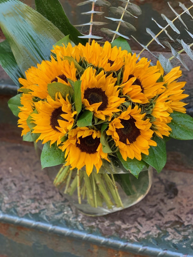 Clusters of bright yellow sunflowers with mixed greenery in an oval clear glass vase